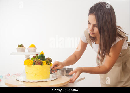 The dark-haired beautiful woman of a confectioner cuts a freshly cooked yellow cake with a knife, decorated with green flowers (roses and cactuses) in Stock Photo