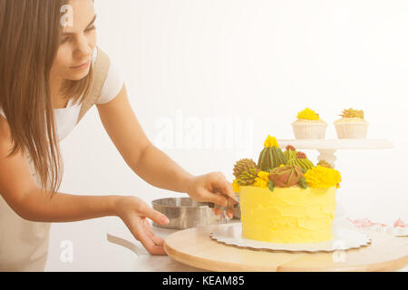 The dark-haired beautiful woman of a confectioner cuts a freshly cooked yellow cake with a knife, decorated with green flowers (roses and cactuses) in Stock Photo