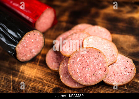 Pile of delicious smoked sausage sliced on a burnt wooden cutting board. Focus on front slice. Red and black sausages blurred in background. Stock Photo