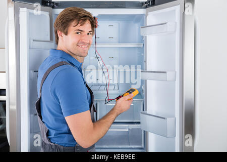 Male Technician In Overall Checking Refrigerator With Multimeter At Home Stock Photo