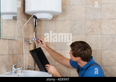 Male Plumber Checking Electric Boiler And Holding Clipboard At Home Stock Photo