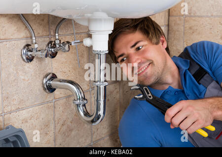 Happy Young Male Plumber Repairing Sink In Bathroom With Wrench Stock Photo