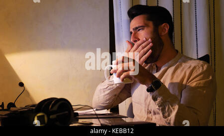 Home worker yawning at computer drinking coffee or tea Stock Photo
