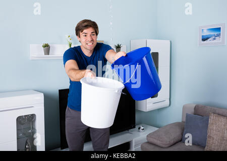 Young Male Worried Man Holding Bucket While Water Droplets Leaking From Ceiling Stock Photo