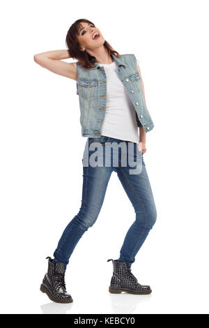 Young woman in jeans vest and black boots is standing legs apart, looking away and laughing. Full length studio shot isolated on white. Stock Photo