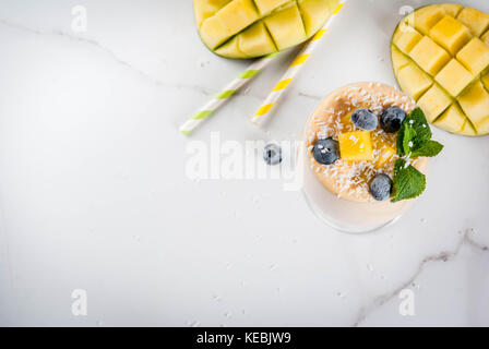 Dietary drink, breakfast. Tropical mango smoothie with fresh pieces of mango, blueberries, coconut and mint leaves. In a glass, on a white marble tabl Stock Photo