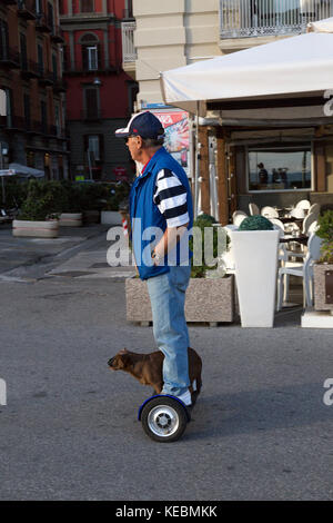 Man with dog riding a hover board along the street in Naples Italy . The dog is wearing sunglasses. Stock Photo