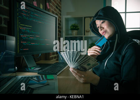 young smiling female hacker looking at banknote thinking how to use programming code stealing credit card information and trading. Stock Photo
