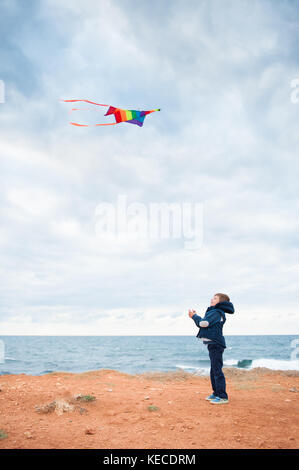 cute small caucasian boy wearing autumn jacket playing flying kite standing on the shore near stormy sea Stock Photo