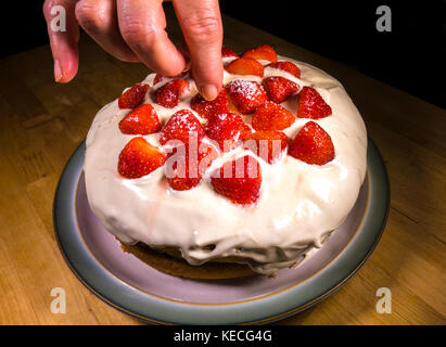 A woman's fingers putting in place the final strawberry on top of a cream topped, sponge birthday cake with strawberries. England, UK. Stock Photo
