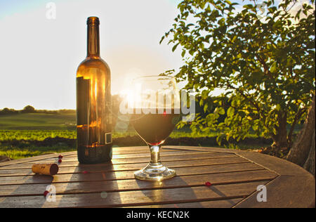 Glass of red wine with open bottle, cork and red berries on wooden table with green fields, bush and sunset in the background Stock Photo