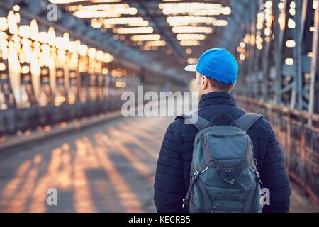 Tourist with backpack walking on the old iron bridge over river at the sunrise. Stock Photo