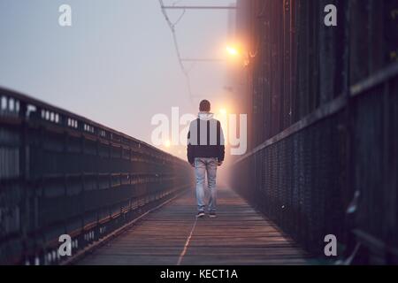 Gloomy weather. Lonely man is walking on the old bridge in mysterious fog. Stock Photo