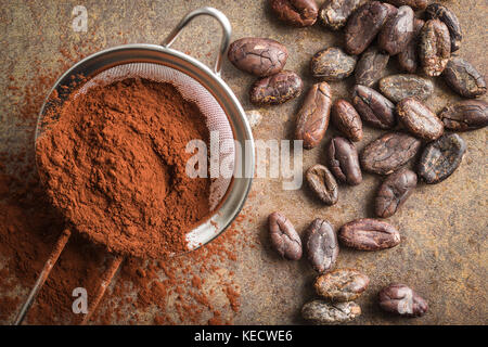 Dark cocoa powder in a sieve and cocoa beans on old kitchen table. Top view. Stock Photo