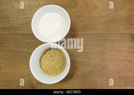 Wheat flour in white ceramic bowl, ramekin, ground toasted sesame seeds in white ceramic bowl, on wooden table, flat lay Stock Photo