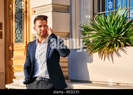A handsome young businessman leaning against the wall at the entrance of a building while talking on his phone in the sunshine Stock Photo