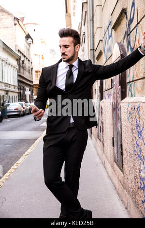 Portrait of a young businessman leaning on the wall, checking his smartphone Stock Photo