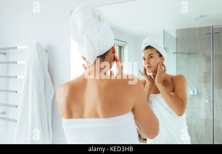 Woman observing her face in mirror in bathroom. Woman in towels wrapped around head and body after bath. Stock Photo