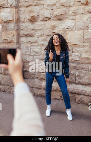 Woman clicking photo of her friend using mobile phone. Joyful woman posing for a photo while her friend uses mobile phone to capture the photo. Stock Photo