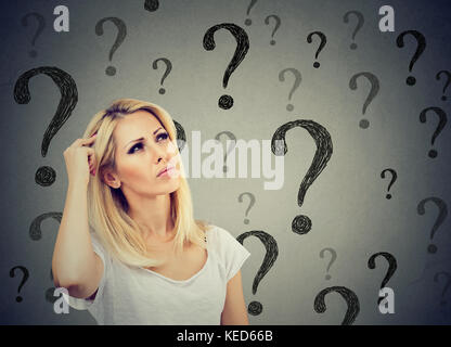 Portrait confused thinking woman bewildered scratching head seeks a solution looking up at many question marks isolated on gray wall background. Human Stock Photo