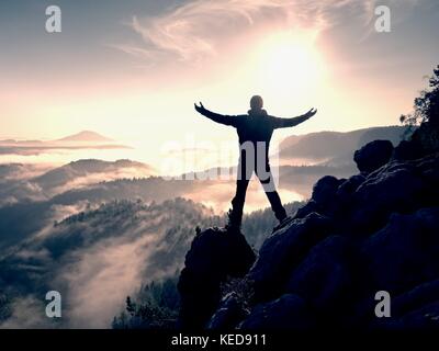 Sunny fall morning. Happy hiker with raised hands in air stand on rock above pine forest. View over misty and foggy morning valley to Sun. Stock Photo