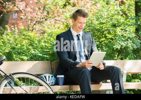 Smiling Male Businessman Sitting On Bench Using Digital Tablet At Park Stock Photo