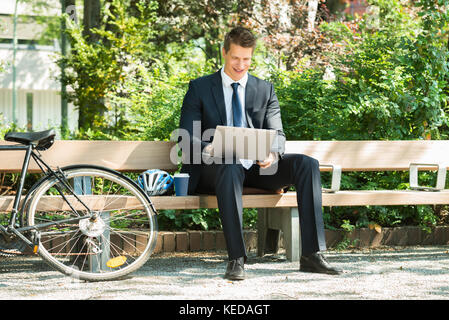 Happy Male Businessman Sitting On Bench Using Laptop At Park Stock Photo