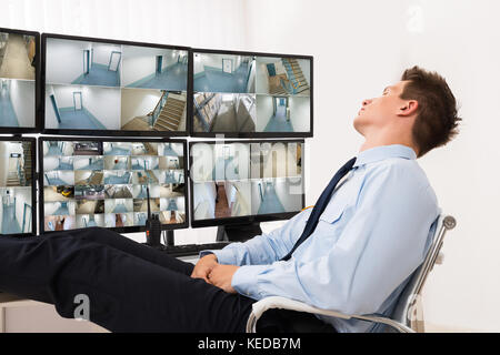 Security Guard Sleeping In Front Of Multiple Computers Showing CCTV Footage Stock Photo