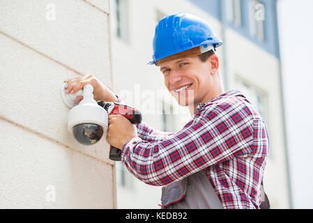 Young Male Technician Installing Camera On Wall Using Electric Cordless Drill Stock Photo