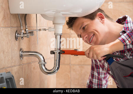 Smiling Male Plumber Fixing Sink Pipe With Adjustable Wrench In Bathroom Stock Photo