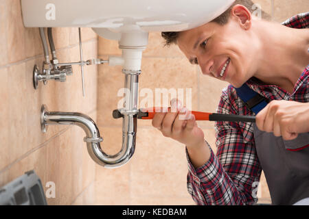 Smiling Male Plumber Fixing Sink Pipe With Adjustable Wrench In Bathroom Stock Photo