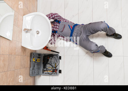 High Angle View Of Male Plumber Lying On Floor Repairing Sink In Bathroom Stock Photo