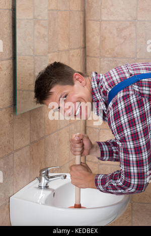 Smiling Young Male Plumber Using Plunger In Clogged Sink In Bathroom Stock Photo