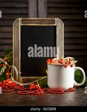 Christmas glowing garland in cup, chalk board, fir branches and rowan berries on dark background Stock Photo