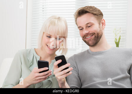 Young Happy Couple Sitting On Couch Using Cellphone Stock Photo