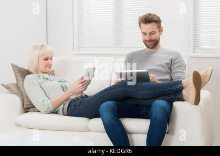 Young Couple Relaxing On Sofa Using Digital Tablets At Home Stock Photo