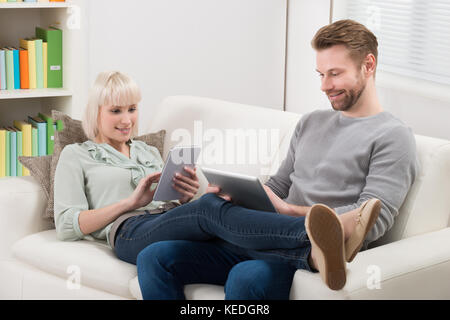 Young Couple Relaxing On Sofa Using Digital Tablets At Home Stock Photo