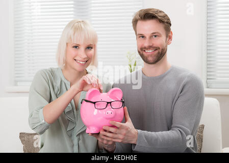 Young Happy Couple Sitting On Sofa Inserting Coin At Piggybank Stock Photo