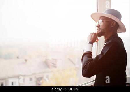 Rich african man looking at panoramic window at his penthouse. Portrait of successful black man in hat indoor. Stock Photo