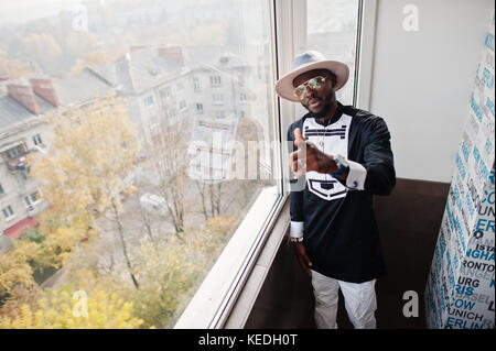 Rich african man looking at panoramic window at his penthouse. Portrait of successful black man in hat indoor. Stock Photo