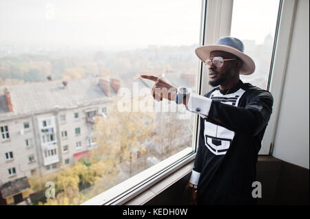 Rich african man looking at panoramic window at his penthouse. Portrait of successful black man in hat indoor. Stock Photo