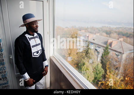 Rich african man looking at panoramic window at his penthouse. Portrait of successful black man in hat indoor. Stock Photo