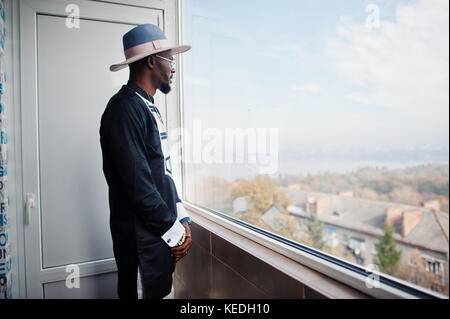 Rich african man looking at panoramic window at his penthouse. Portrait of successful black man in hat indoor. Stock Photo