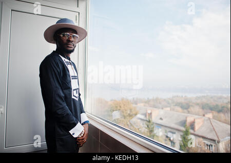 Rich african man looking at panoramic window at his penthouse. Portrait of successful black man in hat indoor. Stock Photo