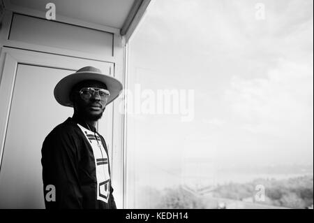 Rich african man looking at panoramic window at his penthouse. Portrait of successful black man in hat indoor. Stock Photo