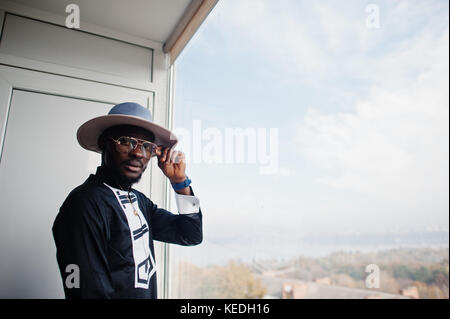 Rich african man looking at panoramic window at his penthouse. Portrait of successful black man in hat indoor. Stock Photo