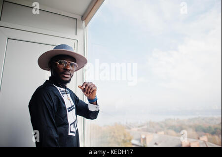 Rich african man looking at panoramic window at his penthouse. Portrait of successful black man in hat indoor. Stock Photo