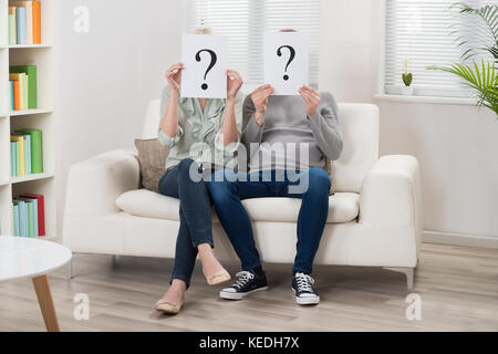 Couple Sitting On Sofa Holding Question Mark Sign In Front Of Face Stock Photo