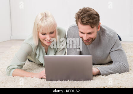 Happy Young Couple Using Laptop Together While Lying On Rug At Home Stock Photo