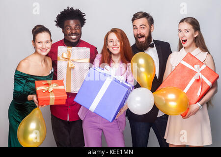 Beautiful people celebrating new year. Holding many gift box, air balloon, looking at camera and toothy smiling. Studio shot on gray background Stock Photo